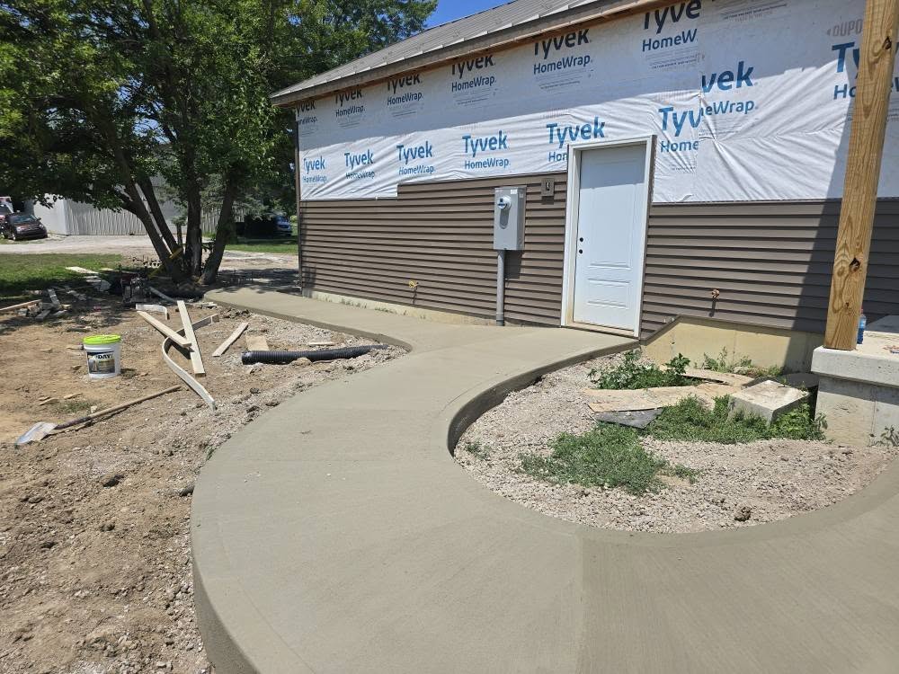 A newly paved concrete walkway curves along the side of a building under construction, wrapped in Tyvek. Tools and a bucket are scattered nearby.