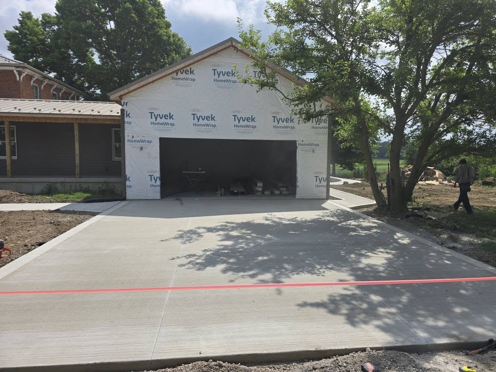 A newly constructed garage with Tyvek wrap and a smooth concrete driveway. A red line stretches across the driveway. Trees and a person in the background.