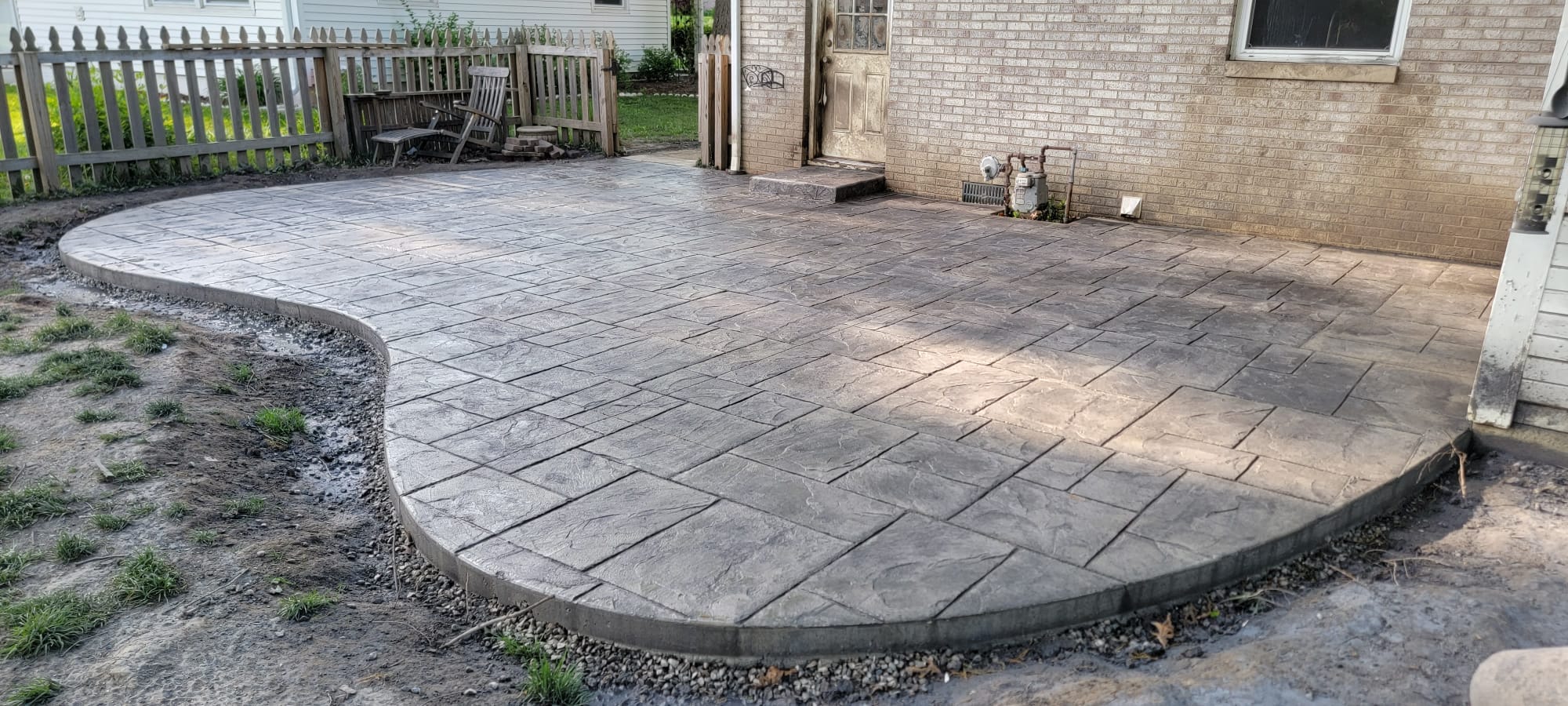 A textured concrete patio with a curved edge beside a brick house, bordered by a wooden fence and garden chairs. The setting is calm, sunny, and welcoming.