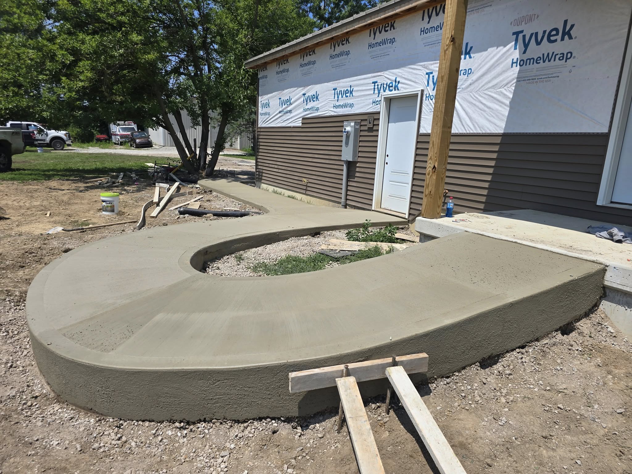 A freshly poured, curved concrete sidewalk leading to a house under renovation. The house is wrapped in Tyvek, with trees and cars nearby.