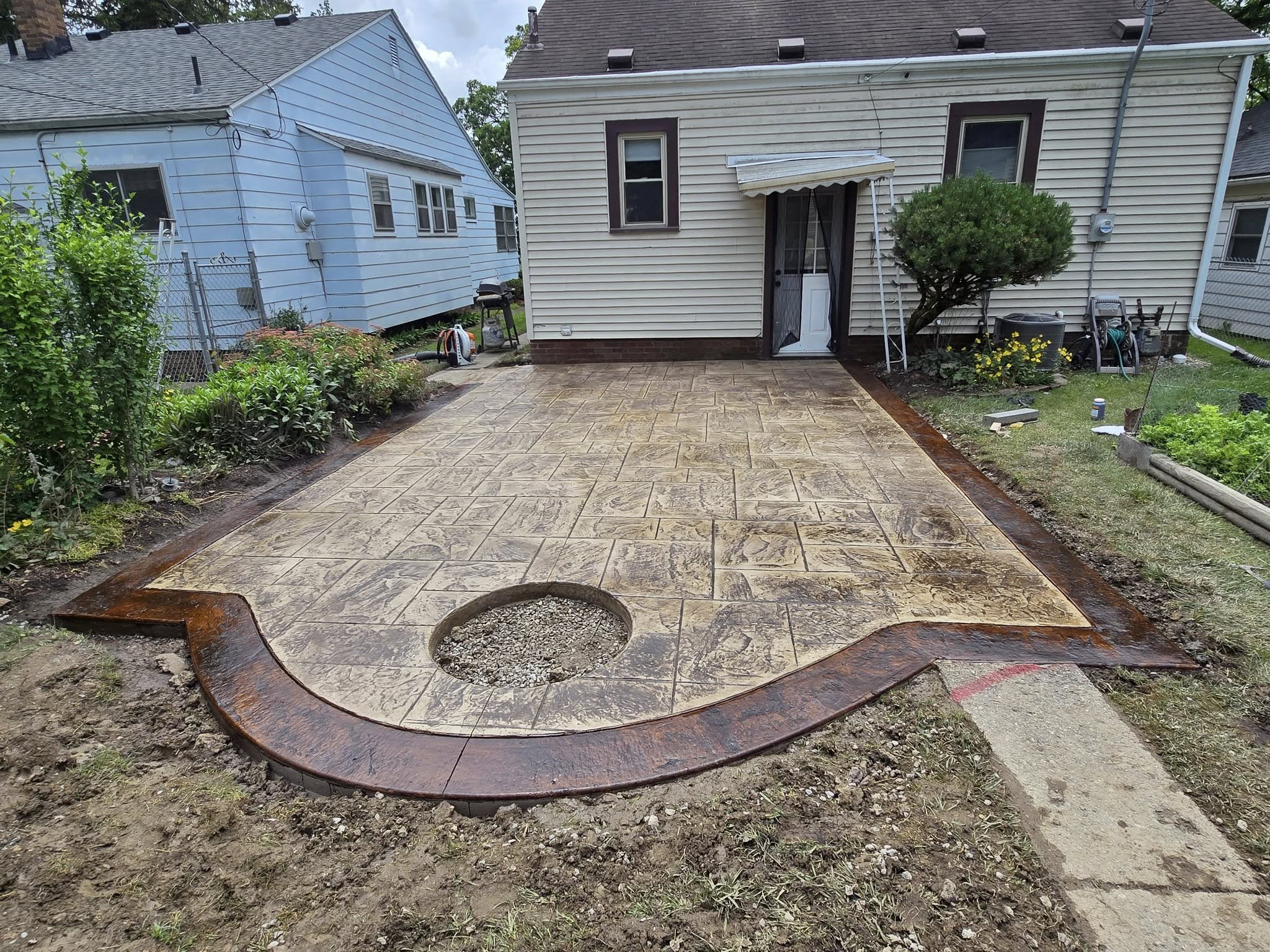 Stamped concrete patio leading to a house with tan siding. A circular hole filled with gravel is in the center. Surrounding area has greenery.