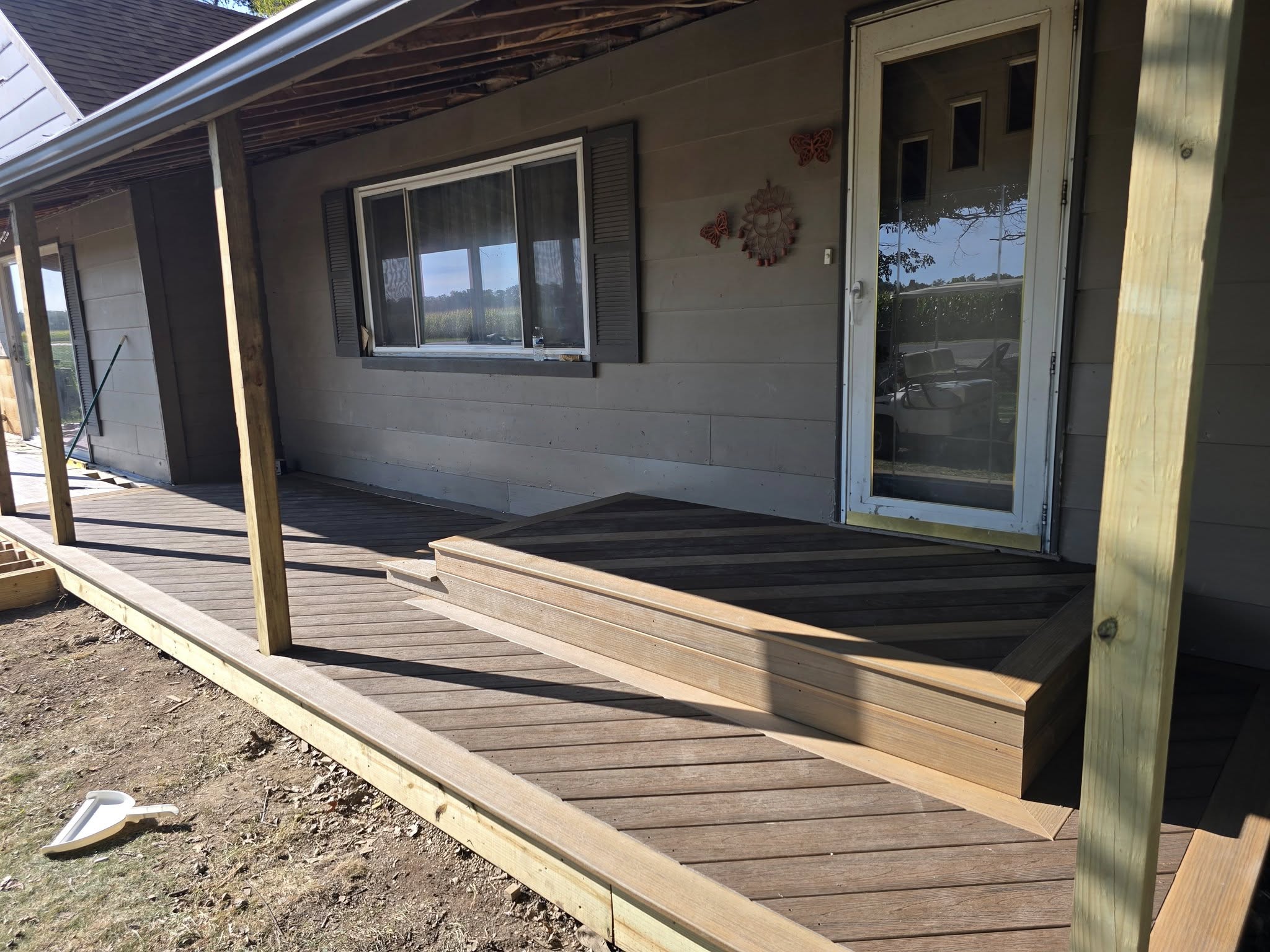 A wooden porch with steps leading to a door, adjacent to a large window, set against a sunny backdrop. The setting is calm and inviting.