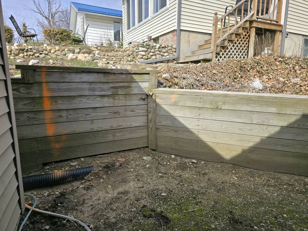 Wooden retaining walls form a corner with visible orange markings. In the background, a house with a blue roof and stone landscaping enhance the earthy scene.