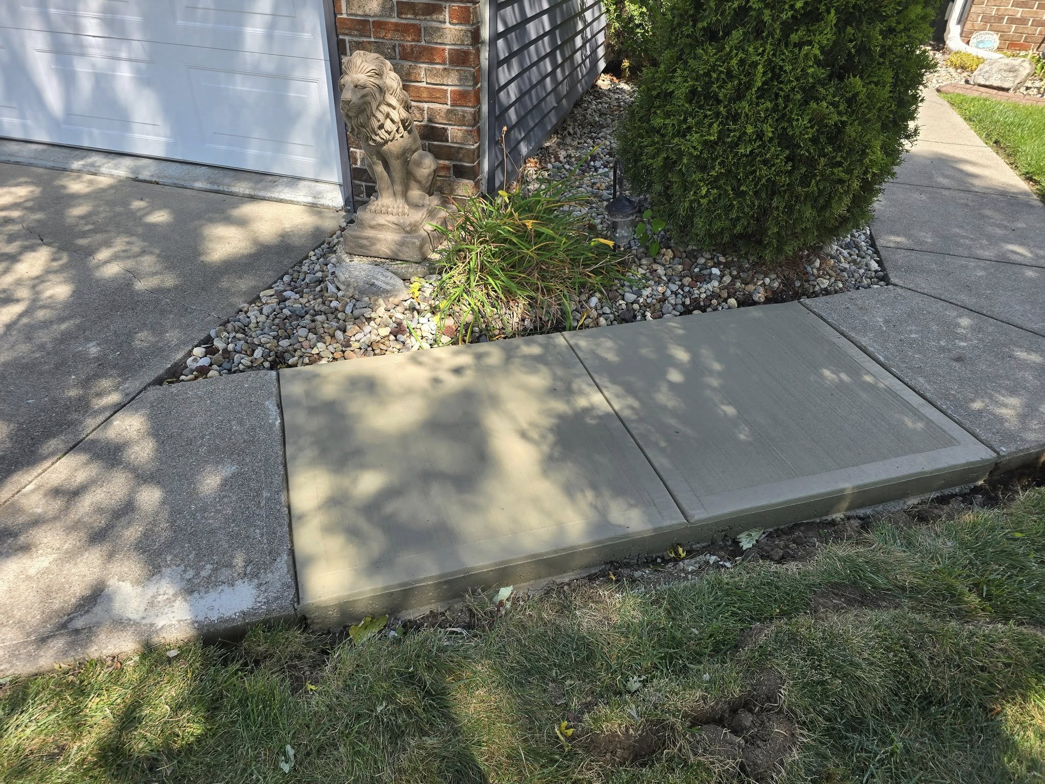 Freshly poured concrete section of a sidewalk, surrounded by a lawn and flower bed with a lion statue. Sunlight creates dappled tree shadows.