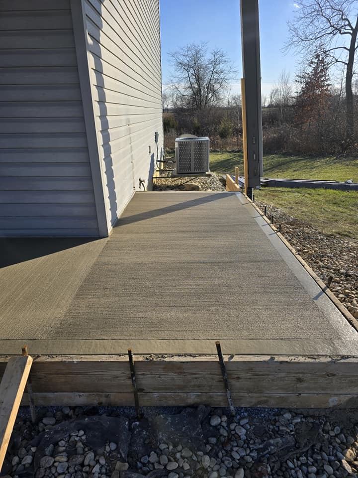Freshly poured concrete patio alongside a beige-sided building, bordered by a grassy yard and trees under a clear blue sky.