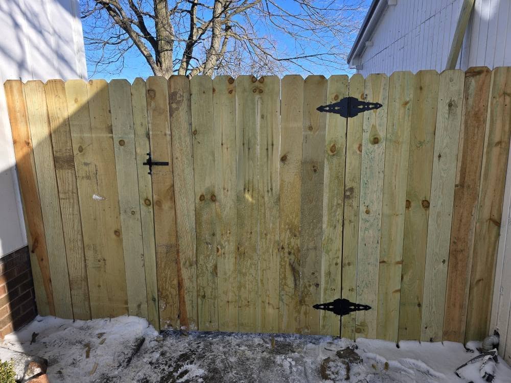 Wooden fence gate with black metal hinges and latch, set against a snowy ground. A leafless tree and blue sky are visible in the background.