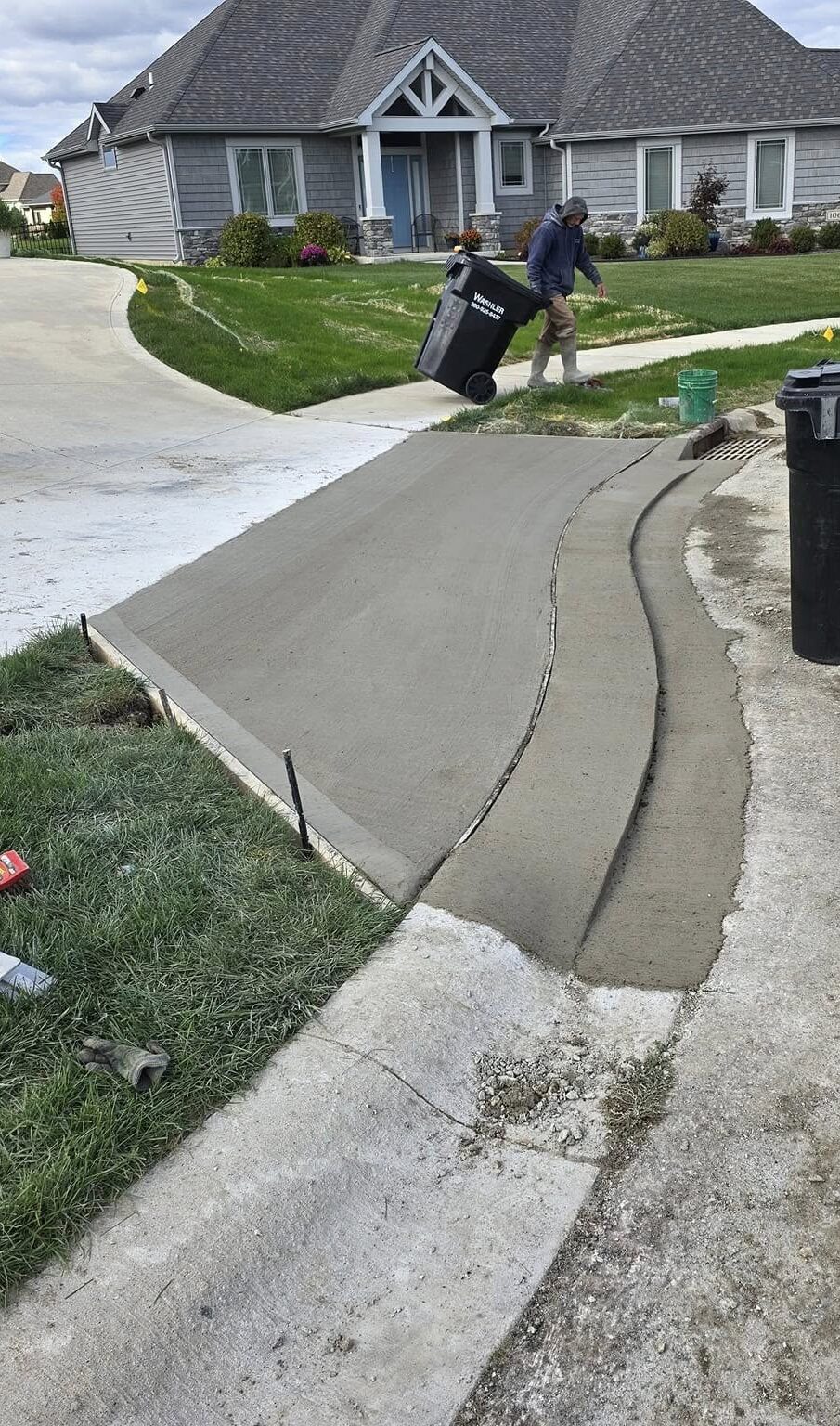 Wet concrete driveway with two steep steps leading to a suburban home. A person is wheeling a trash bin nearby on lush green grass, under a cloudy sky.